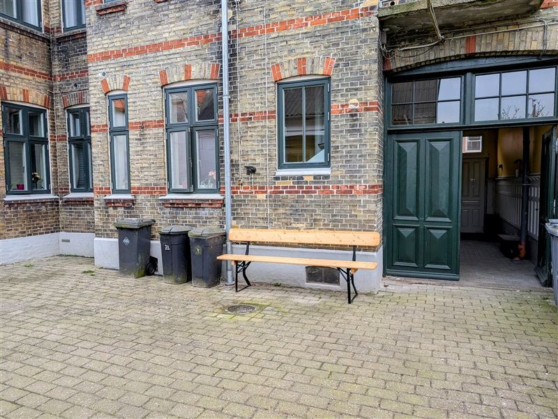 En træbænk og 3 skraldespande udenfor en rødmalet bygning med vinduer og en grøn dør