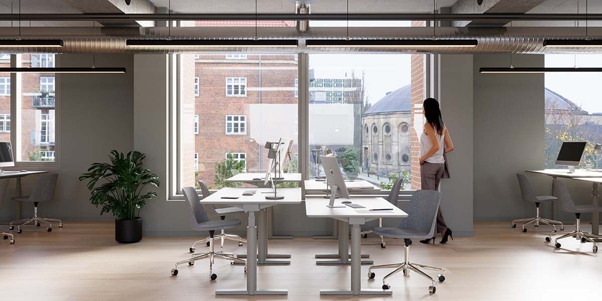 Moderne kontor med lyse lofter, høje vinduer og trægulve. Bord og stol samt plante i hjørnet. Kvinde i stående stilling og