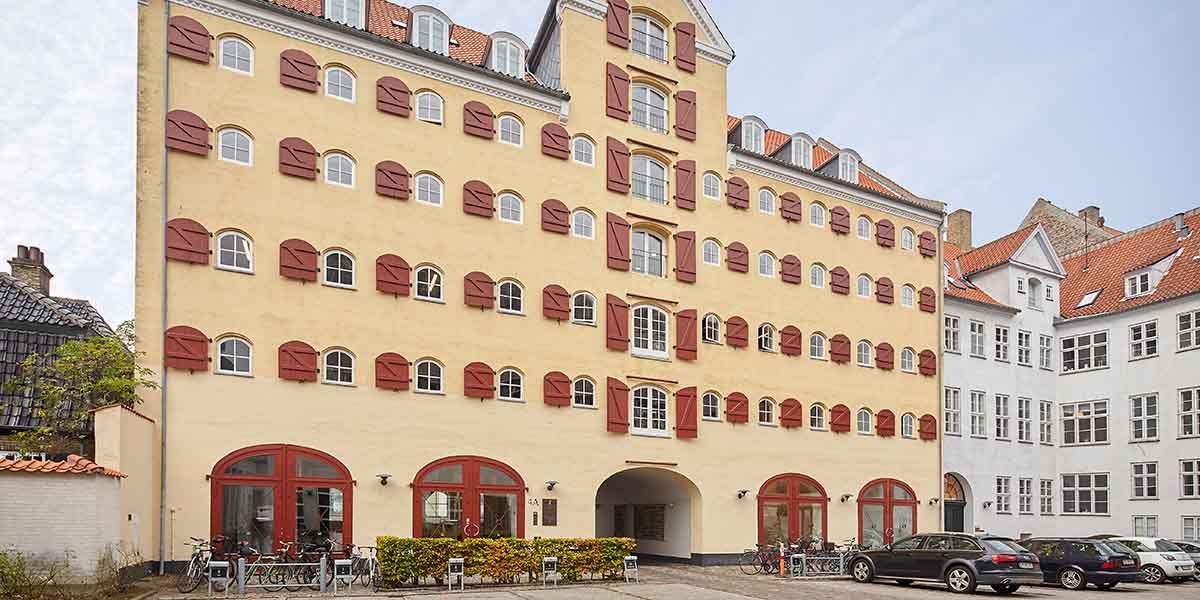 Det Gule Pakhus på Strandgade med røde skodder og gule vægge har flere biler parkeret foran, samt cykler og grønne buske.