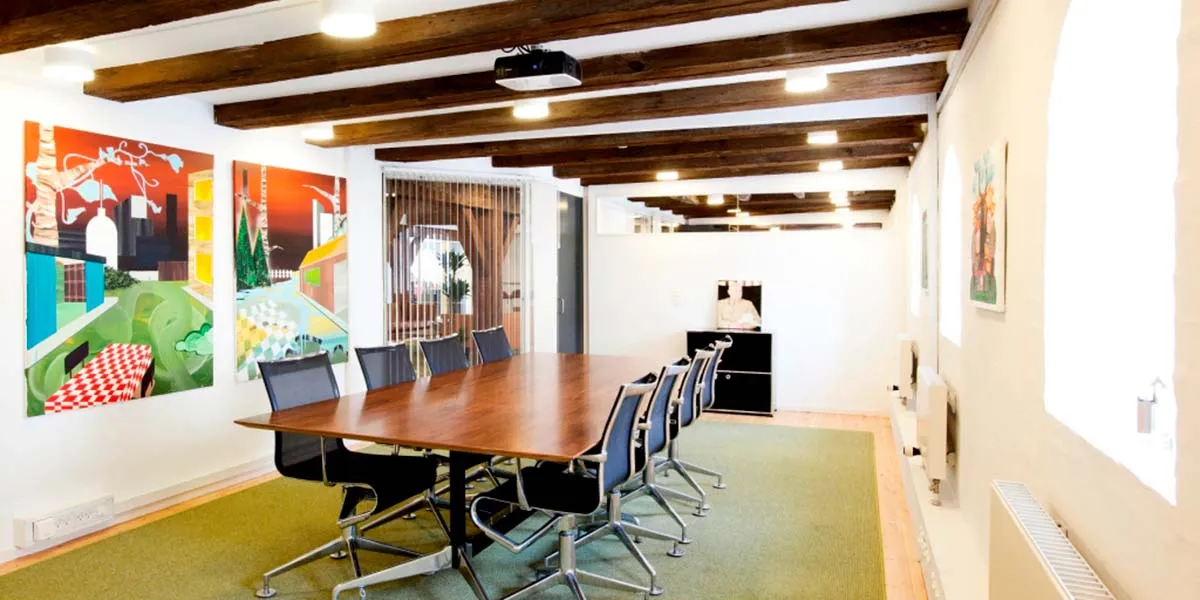 Et kontor med lyse trælofter og malerier på væggen, en lang brun bord med flere stolpe og en projektor.