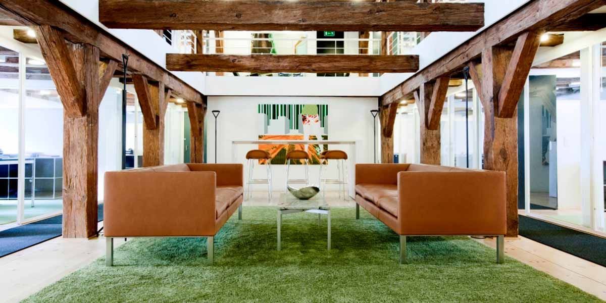 To lædersofaer med metalben samt en stor, grøn gulvtæppe i en stor stue med store træbjælker på loftet og glasvinduer