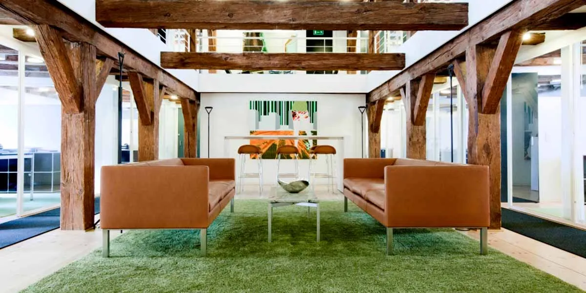 To lædersofaer med metalben samt en stor, grøn gulvtæppe i en stor stue med store træbjælker på loftet og glasvinduer