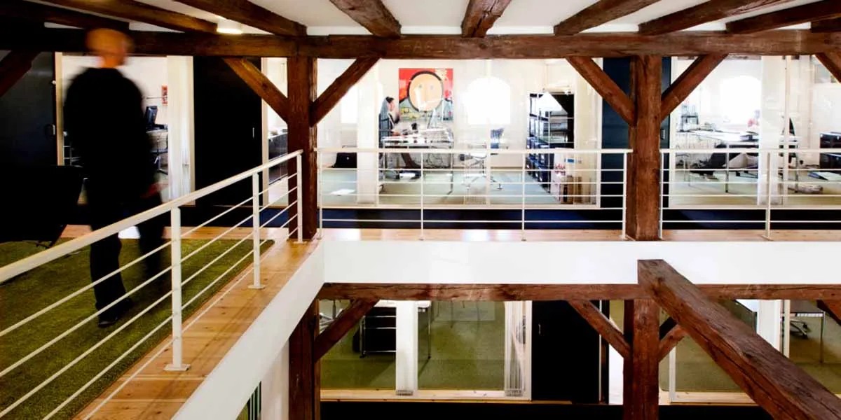 Inde i et gult pakhusejendom er der en loftsterrasse med træbjælker og et hvidt metalgevær. Der er et grønt gulvtæppe og en