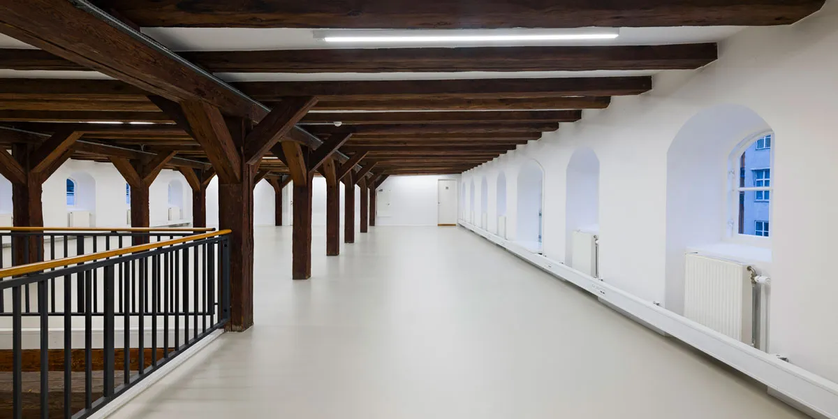 Lang, ubeboet korridor med hvide vægge, lysegul gulv, højt loft med træbjælker og bueformede vinduer