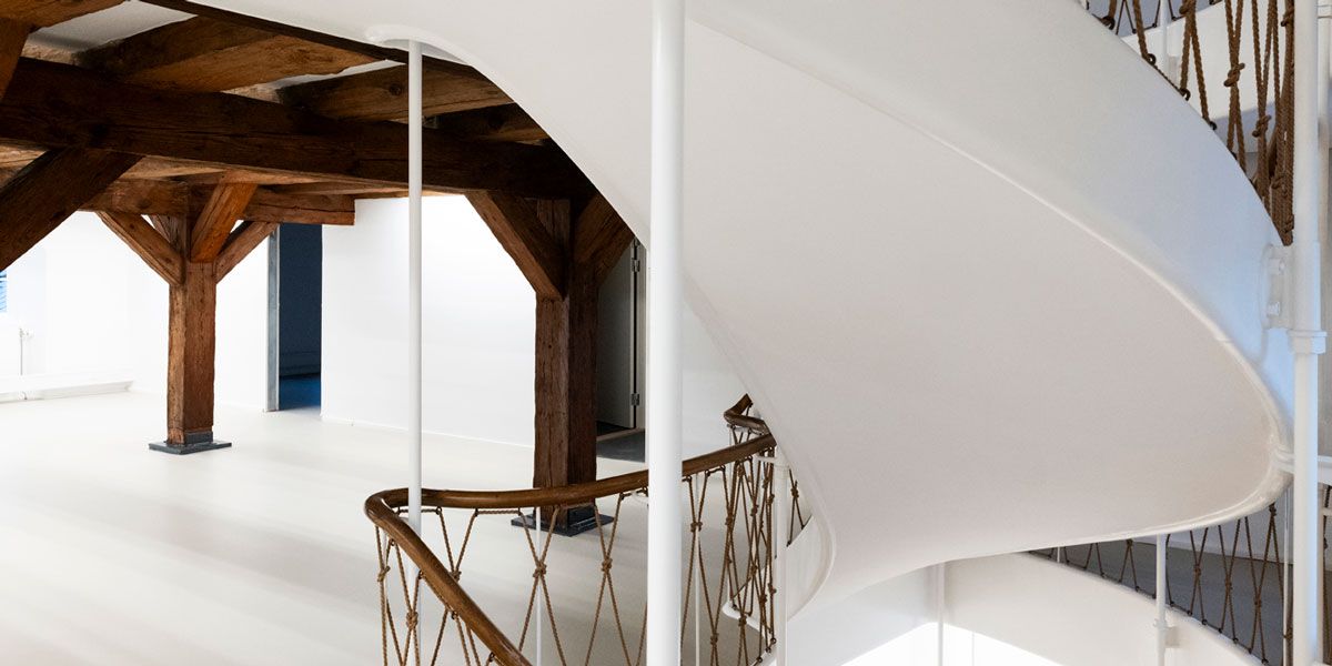 En lyse, smal, hvid snortrappe med brun metalgevær og lyse træbalk i en renoveret bygning med fredet udseende