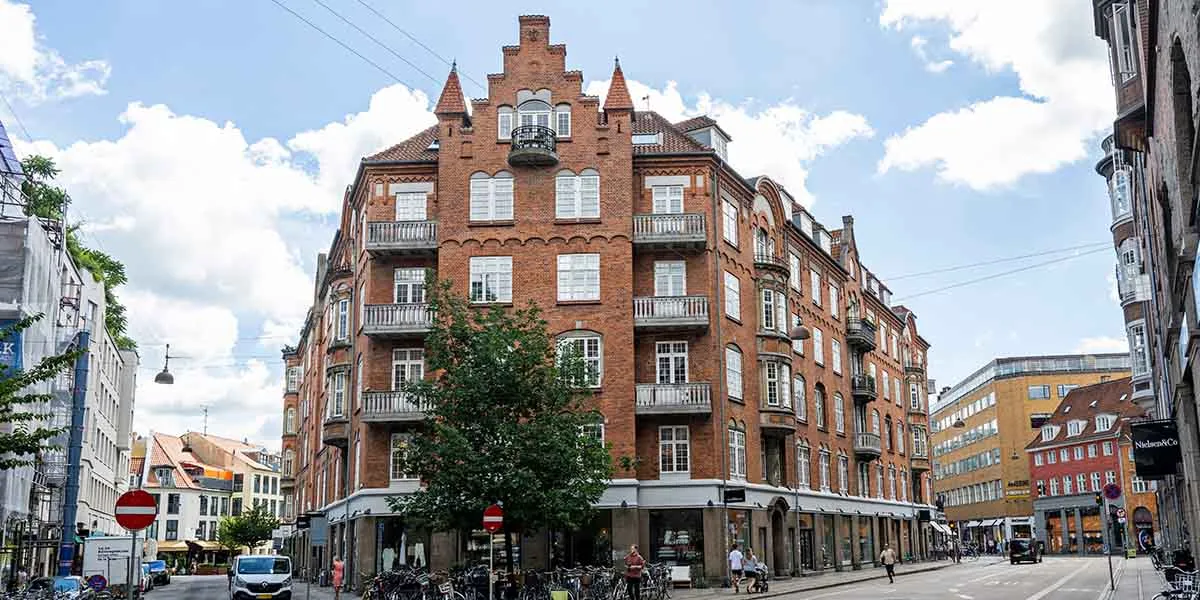 En stor, gammel bygning med røde mursten og flere balkoner og vinduer i Københavns centrum. Mennesker går på fortovet, og