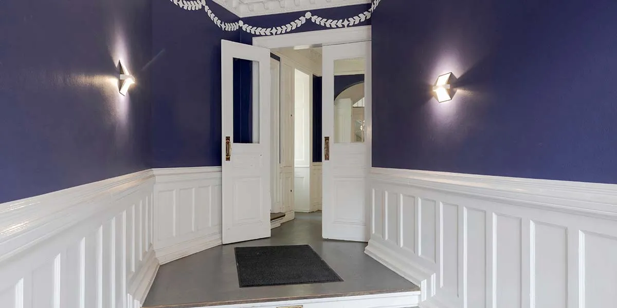 Elegant korridor med mørkeblå vægge, hvide paneler og lysekroner i hjørnerne, åbne hvide døre og sort tæppe på gulvet
