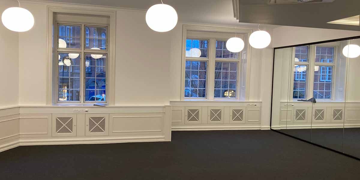 Et lukket kontorrum med hvide vægge, hvide paneler og tre vinduer. Loftet er hvidt med fire runde, hvide loftslampe. Gulvet