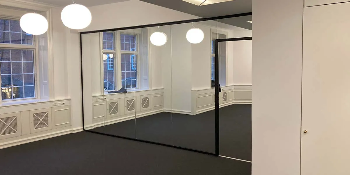 Glasdøre med svære, slanke profiler og gulvbelægning i sort. Vægge og loft er hvide. På gulvet er der refleksioner fra