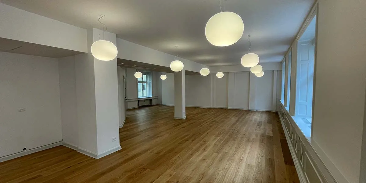 En stor, åben, ledig kontorlokal med lyse loftsvinduer og lyse runde loftslamper, hvide vægge og lyse gulvbelægning samt