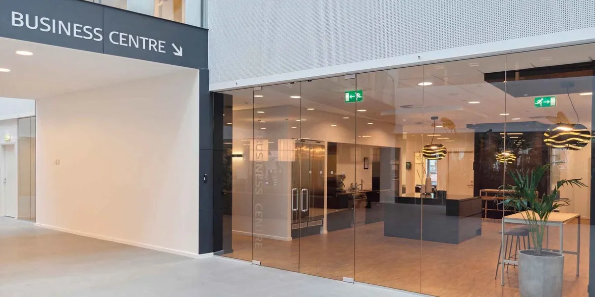 Moderne indgangsparti med glasdøre og skilte, der læser "Business Centre" samt en plante i en stor grå potte på en hvid