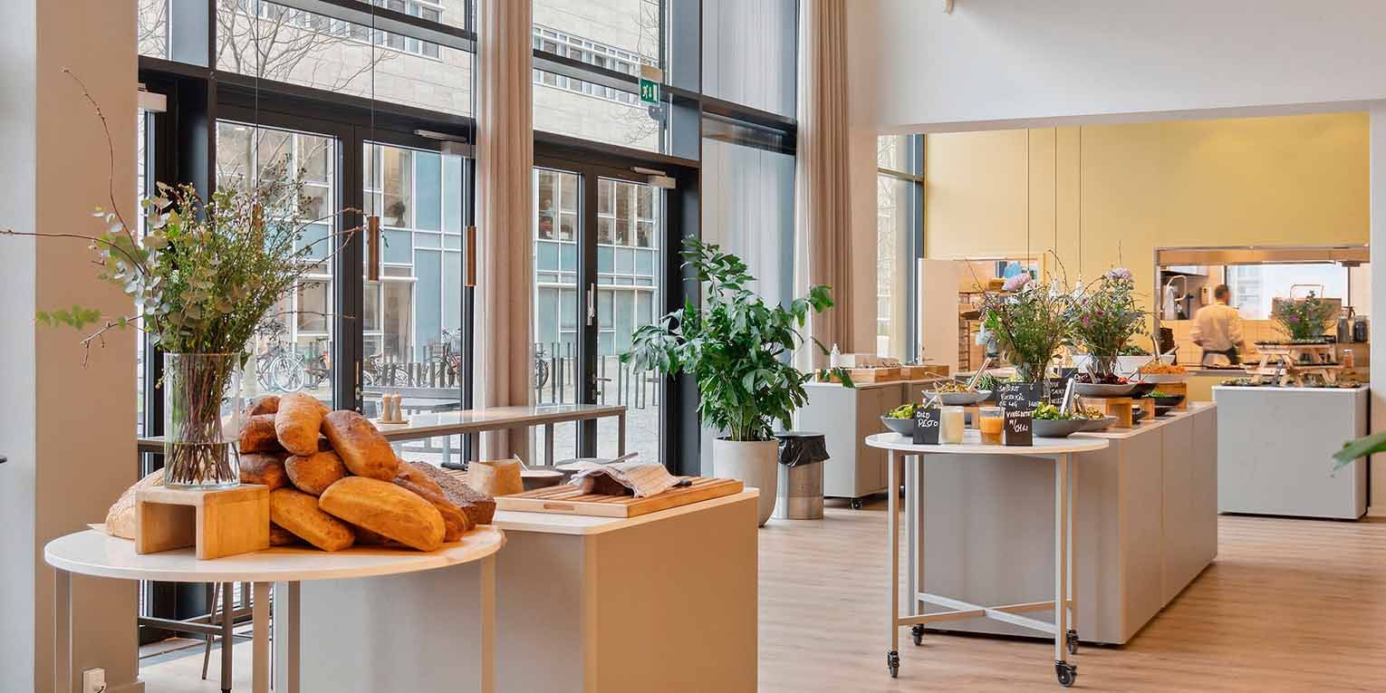 En moderne kontorlounge med trægulve, store vinduer, bord med småplanter og slik, og træskåret bagværk