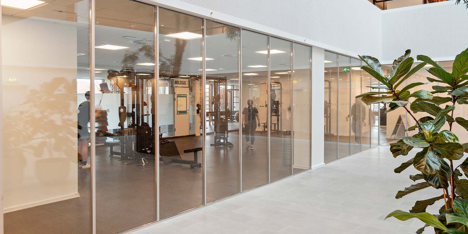 Moderne indretning med store glasvinduer og fitnessapparater i holdesalen. Blomster i hjørnet og en række lysepunkter på