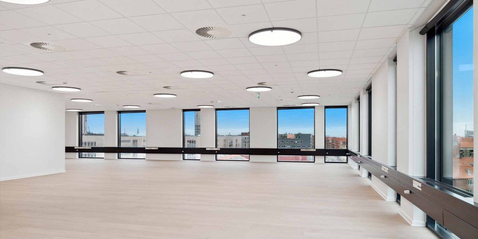 Stor, lyse stue med højloft, lysegul gulvbelægning og store vinduer med udsigt over byen.