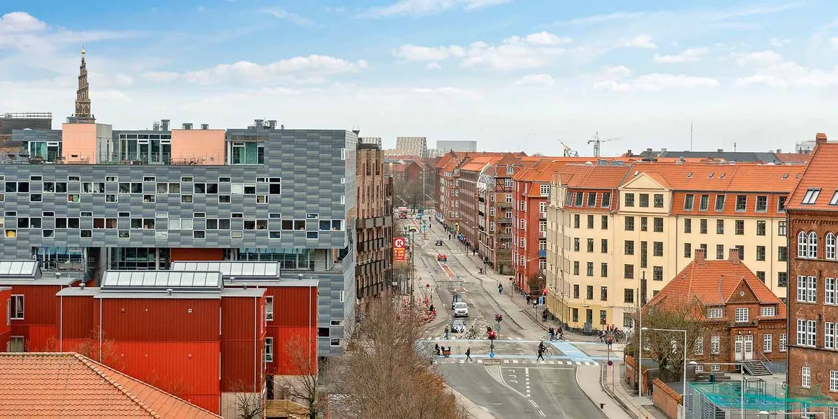 Gadebillede med bygninger og røde tag. I baggrunden er der en bygning med spiretårn og en kran. Vejen er tosporet og har gå-