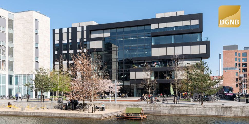 Moderne bygning med glasvinduer og terrasser, omgivet af grønne træer og busser. Folk går forbi, og der er en vandkant med