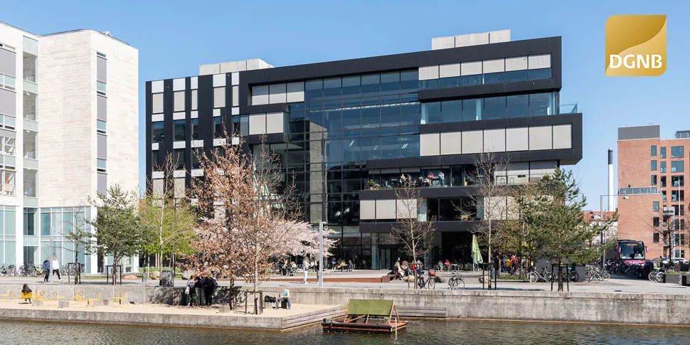 Moderne bygning med glasvinduer og terrasser, omgivet af grønne træer og busser. Folk går forbi, og der er en vandkant med