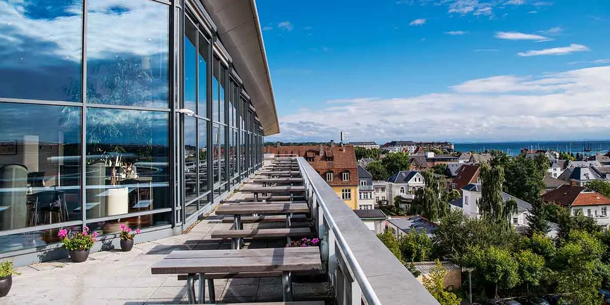 Panoramaudsigt over byens huse og grønne træer, udvendigt fra restaurantens terrasse med store glasruder, bord og blomster.