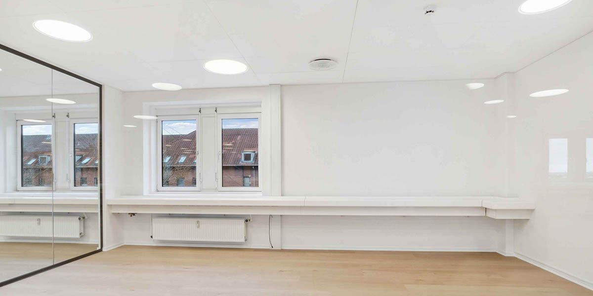 Et moderne rum med hvide vægge, trægulv og store spejle samt radiatorer og to vinduer med udsigt til et hus og en bygning