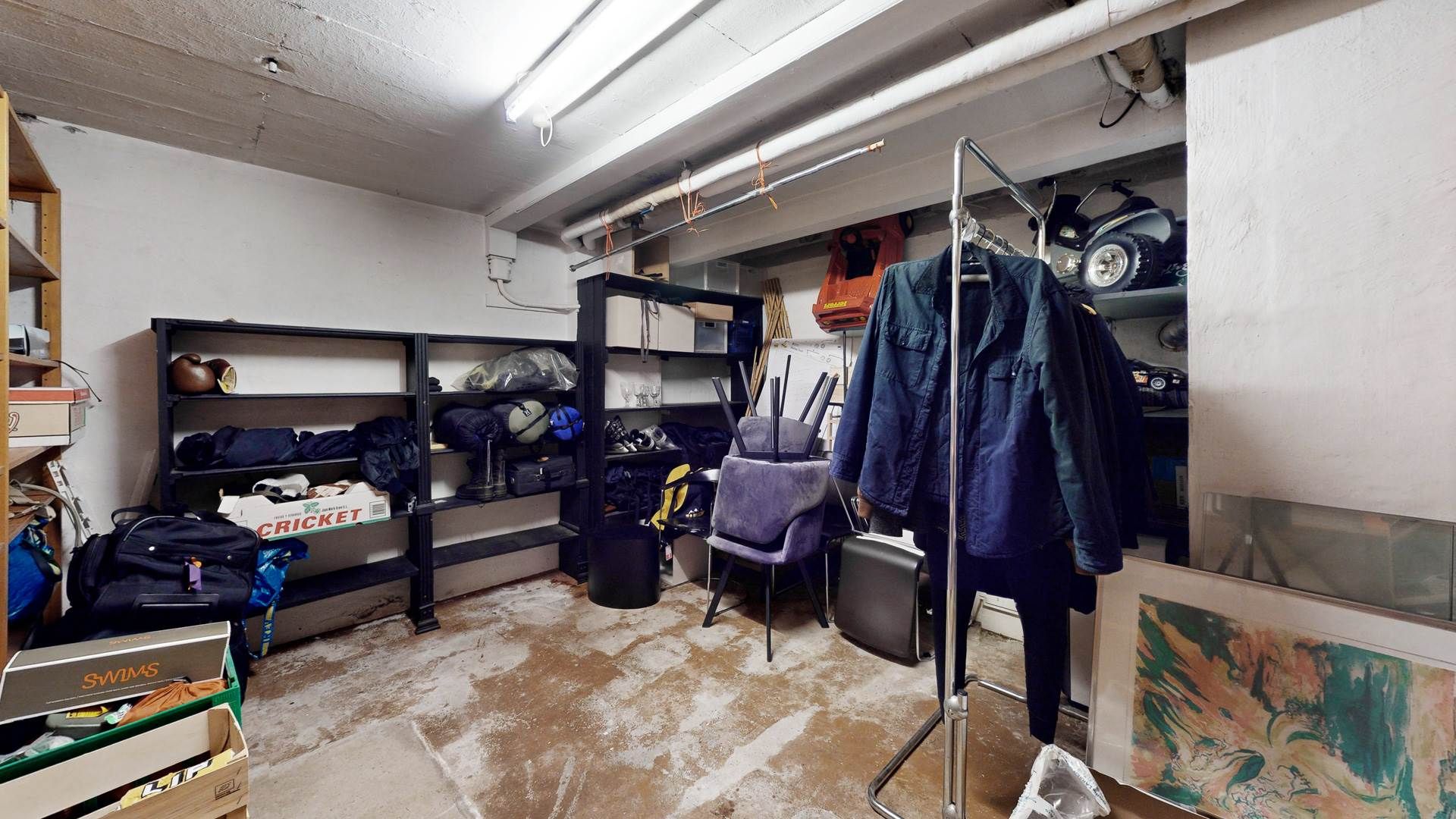 Garage med lagerhylder, tøj, rygsække, senge, stol, tøjstativ, smykker, malerier og en søjle med rør og ledninger