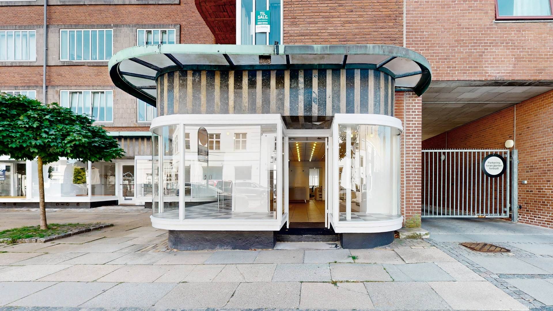 Glaskiosk med runde vinduer og tynde hvide rammer foran en stor bygning med rød mursten og vinduer