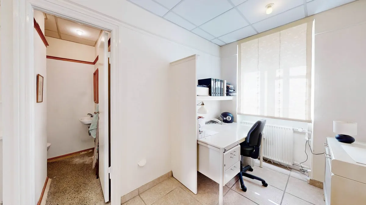 Et hvidt kontorlokale med skrivebord, højtalere, lampe og biblioteksskabe og en smal korridor med toilet og skab.