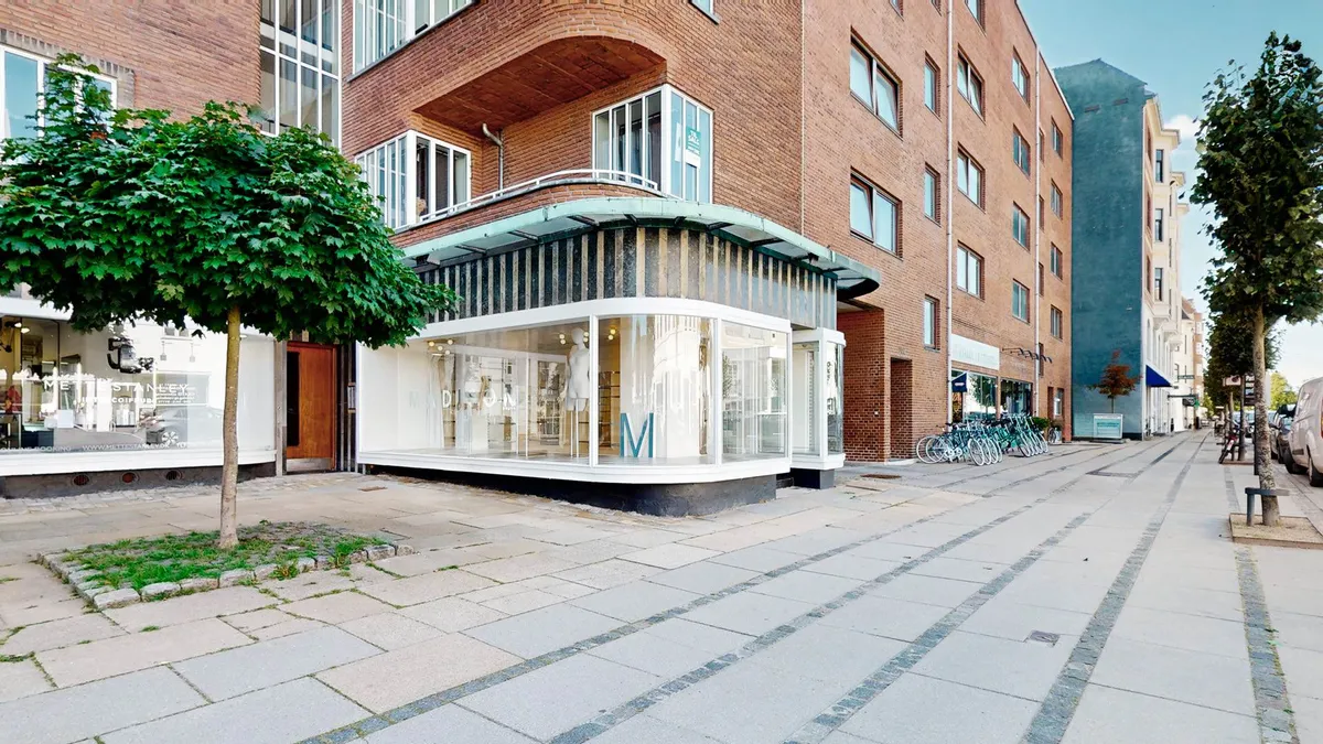 Moderne bygning med glasvinduer og en facade med bogstavet M på forsiden, sammen med et træ på fortovet og cykler parkeret