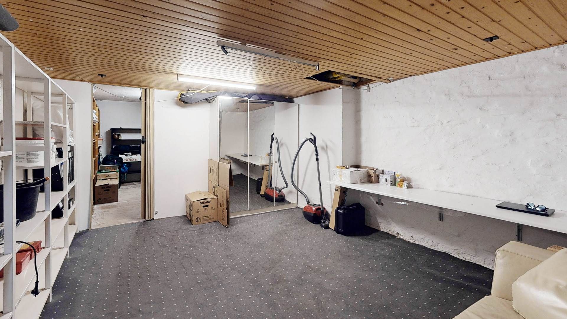 En lille stue med hvide vægge og loft, hvor der er placeret diverse ting, herunder støvsuger, skraldespand og bokse