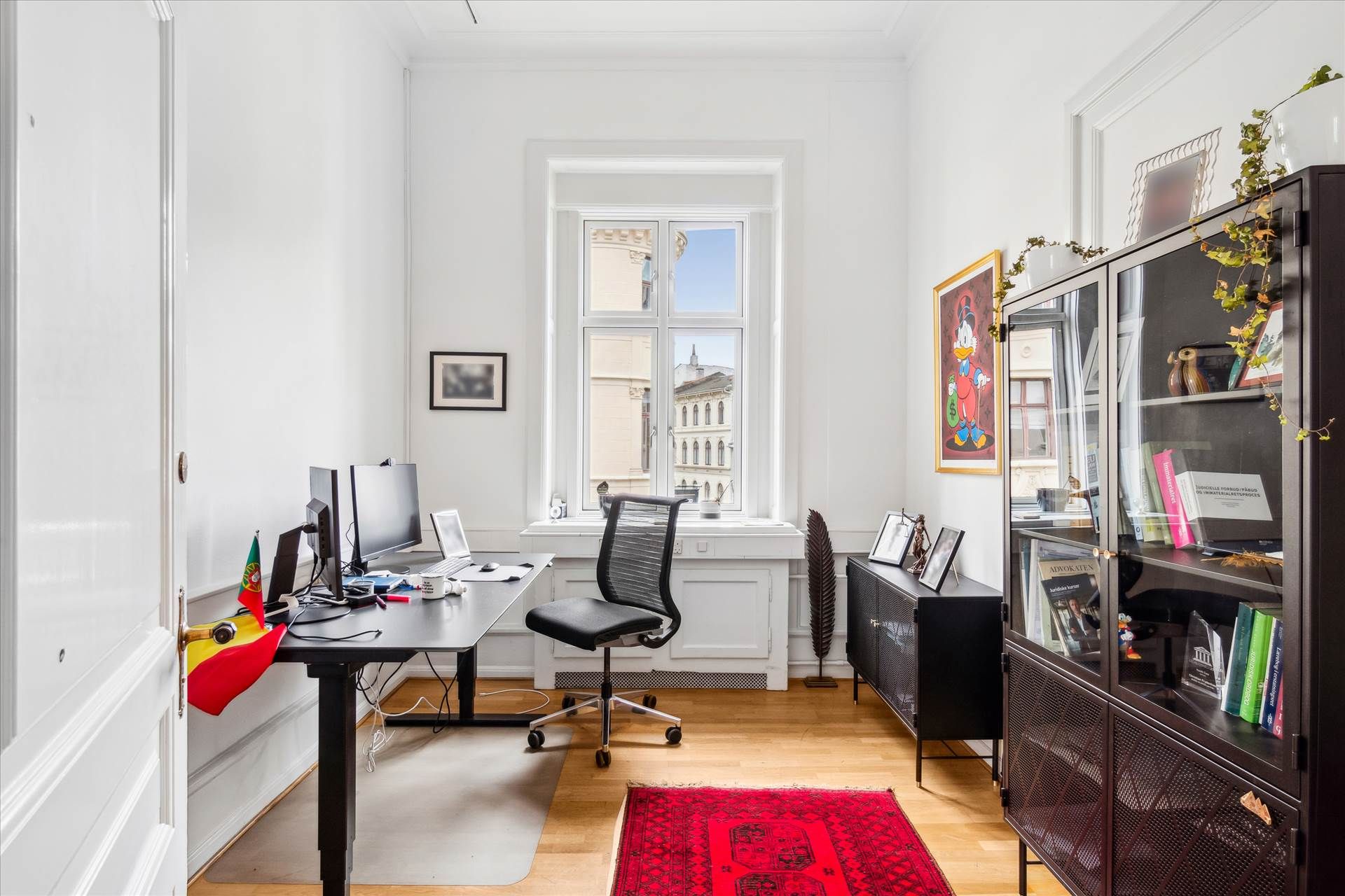 Kontor med højt til loftet, moderne indretning og møbler, gulvtæppe, skrivebord med monitorer, stol, flag og diverse