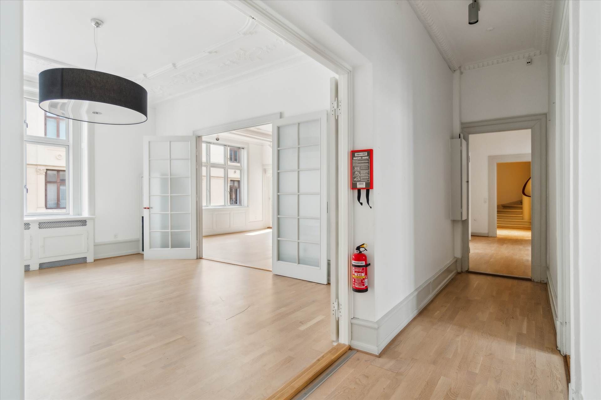 Herskabeligt kontor med højloftede rum og lyse gulve. Et rødt ildslukker og et simpel hængelampe. Adgang til flere rum med