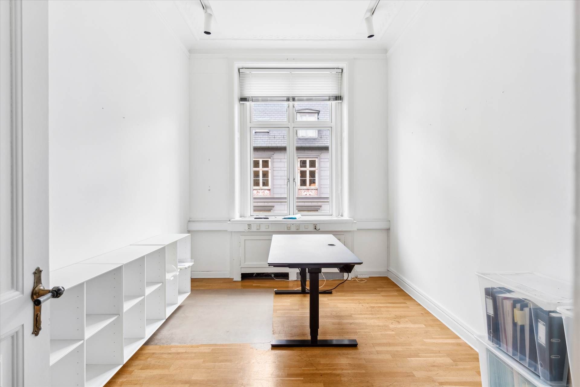 Kontor med højloft og lyse trægulve, store vinduer med gardiner og to høje hvide hylder.