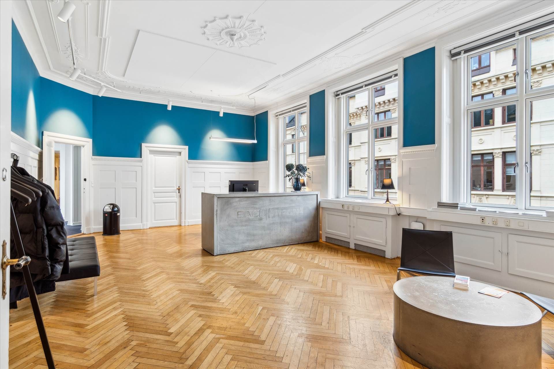 Elegant herskabeligt kontor med massiv trægulv og hvide vægge. Et stort skrivebord, høj vinduesfront og mørke læderstole.