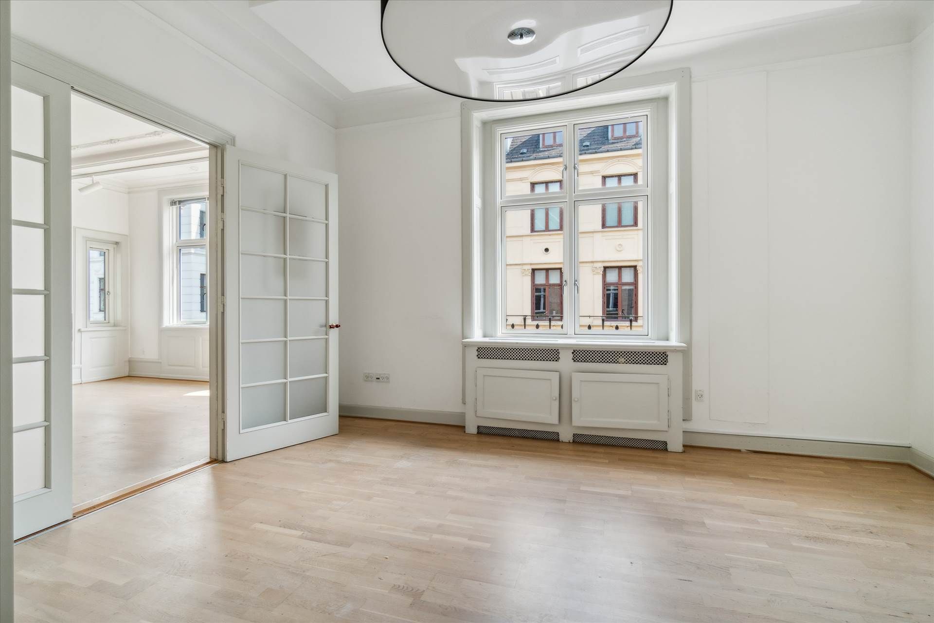 Lukket hvidt glasdør fører til et rum med højt til loftet og lyse gulv. Vindue med balkon og udsigt til et bygning i