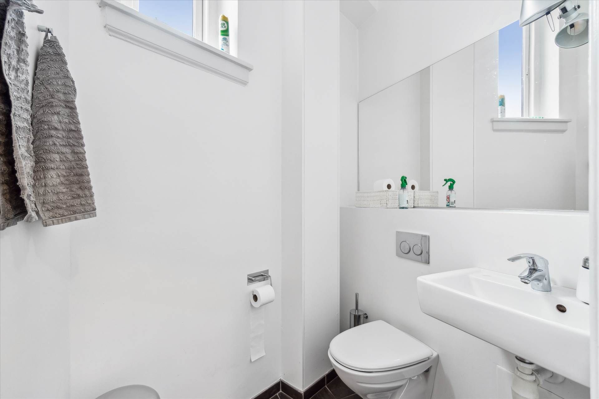 Et moderne badeværelse med hvidt gulv og vægge, en hvidt toilet med låg og en hvidt vask med en sølvfarvet vandhane og to