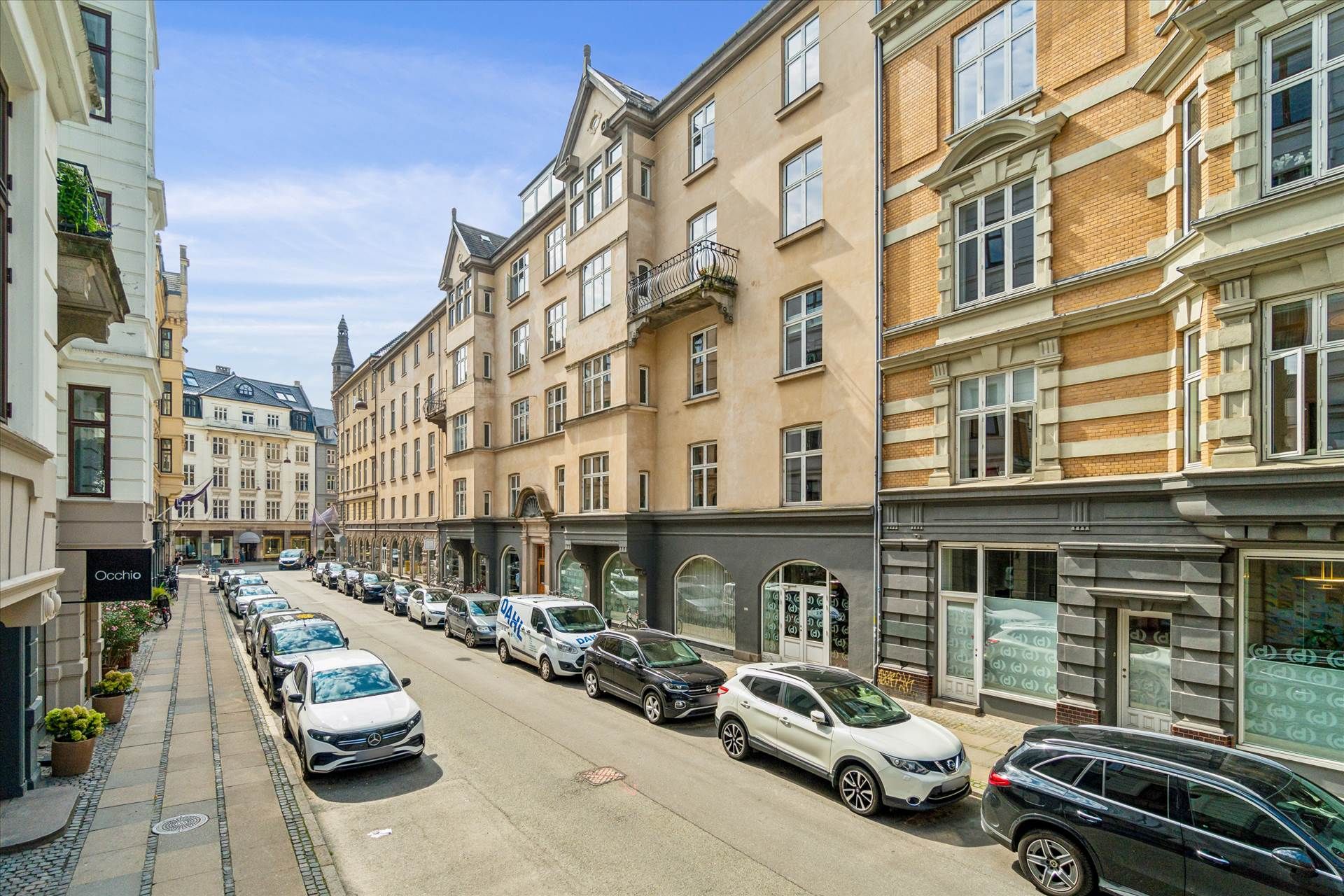 Bredgade med parkerede biler og bygninger med vinduer, balkoner og indgange, samt nogle plantekasser og skilte