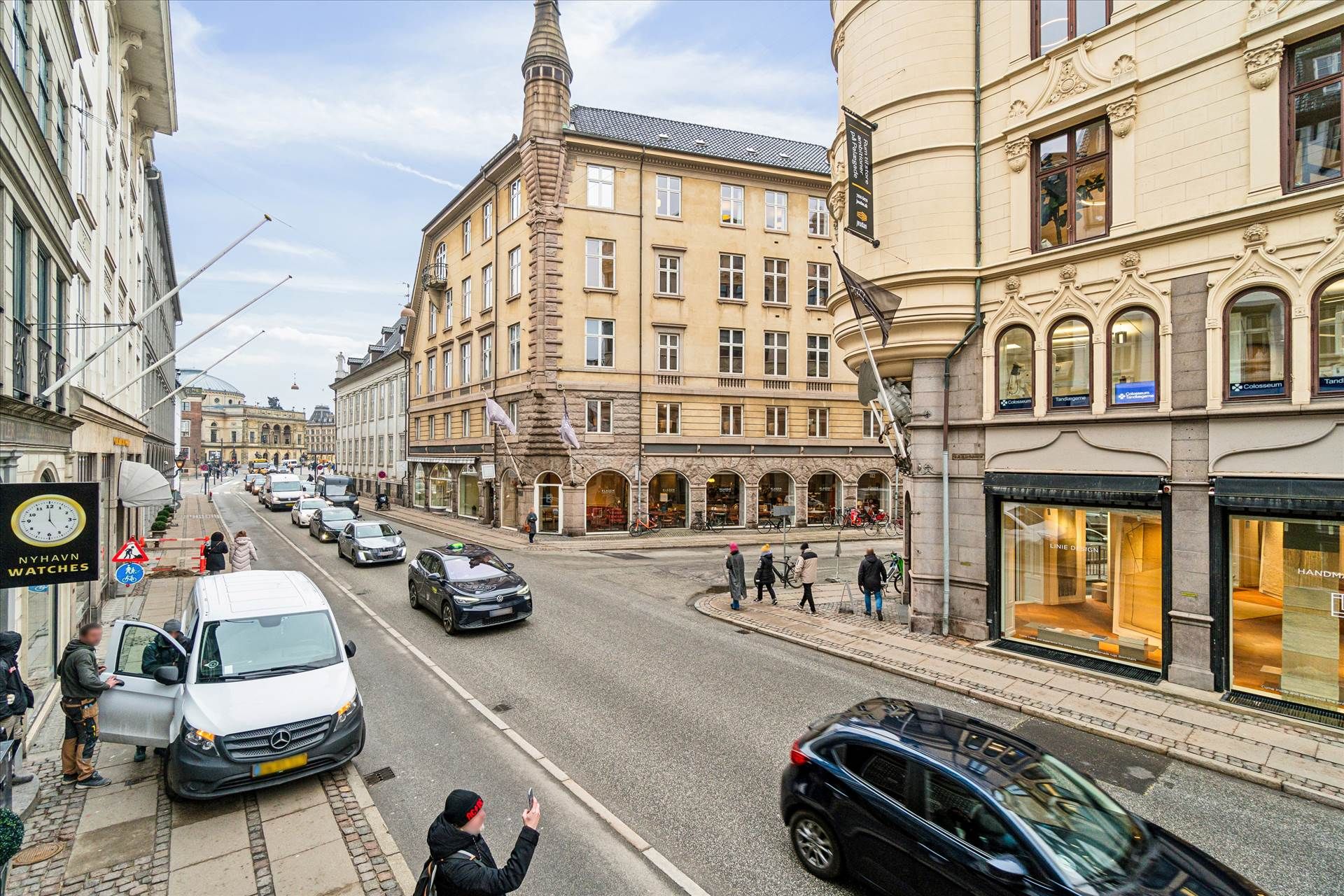 Personer går på gågaden i København med biler og lastbiler, mens en person holder op for at tage et billede