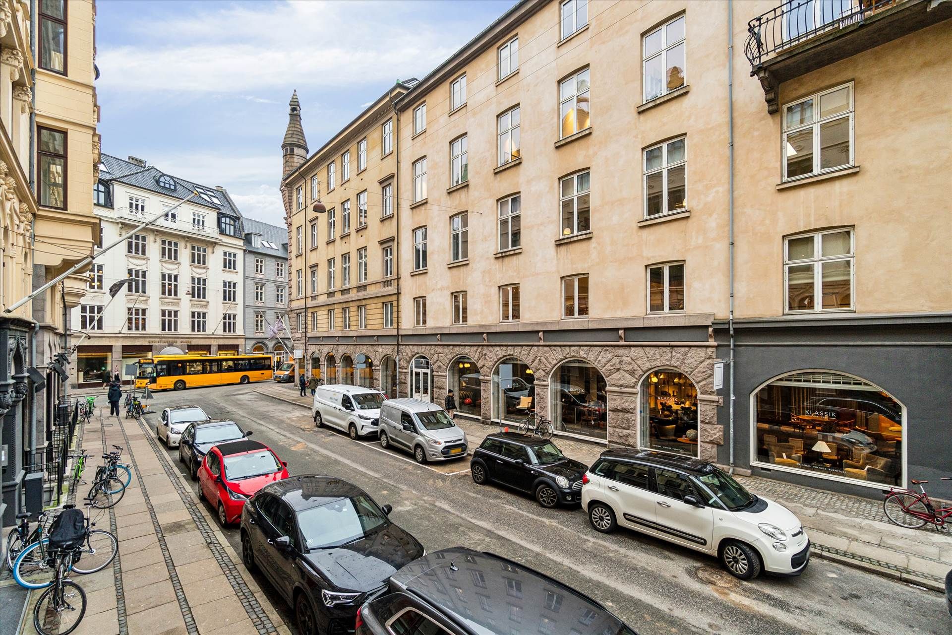 En lang række biler parkeret langs gaden med cykler lænet mod bygningen og en bus i baggrunden