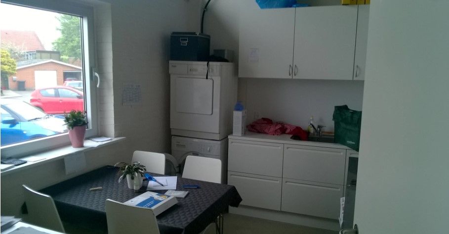 Et teknisk rum med tøjvask, en bordet med en blomst og flere papirer samt hvide køkkenskabe og stole