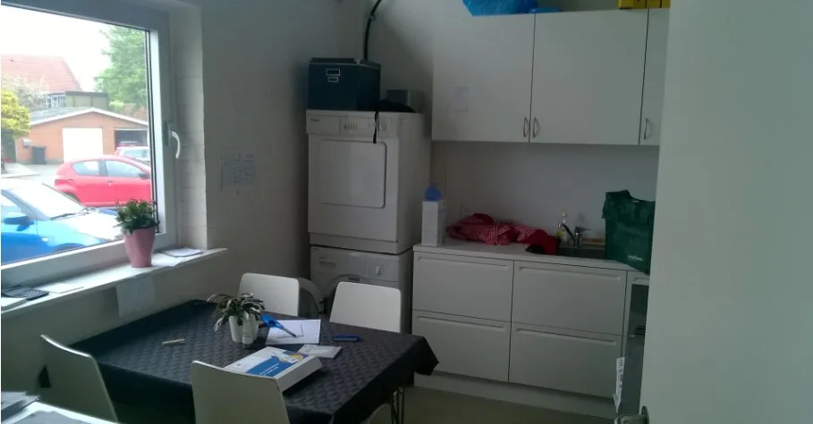 Et teknisk rum med tøjvask, en bordet med en blomst og flere papirer samt hvide køkkenskabe og stole