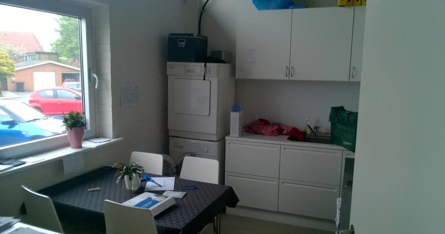 Et moderne køkken med hvide skabsfronter, en bord og stole, en tørretumbler og en vaskemaskine samt en pant i vinduet