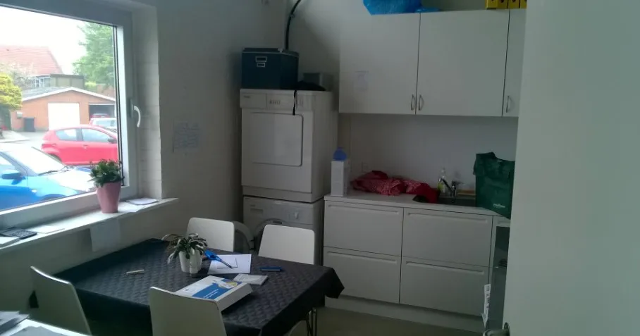 Et moderne køkken med hvide skabsfronter, en bord og stole, en tørretumbler og en vaskemaskine samt en pant i vinduet