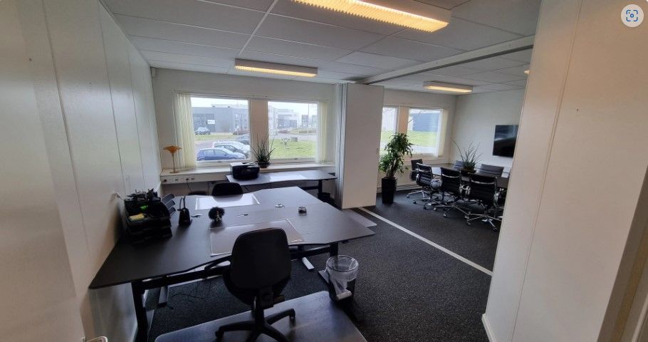 Kontorlokale med lyse loftslampe, bord, stole, plante og fjernsyn i hvid stue med vinduer og parkeringsplads udenfor