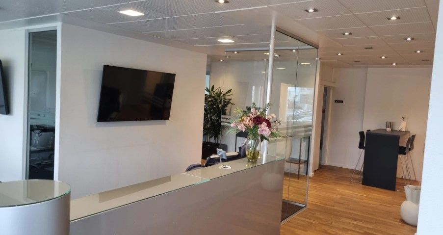 Kontorlokale med masser af lys og glasvægge, og en tv-skærm på væggen, en blomstervase med røde blomster, og en bord med en