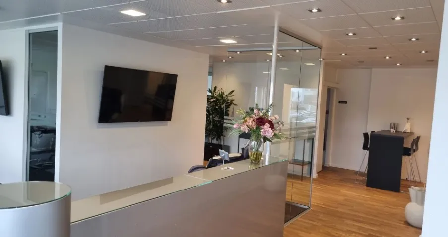 Kontorlokale med masser af lys og glasvægge, og en tv-skærm på væggen, en blomstervase med røde blomster, og en bord med en