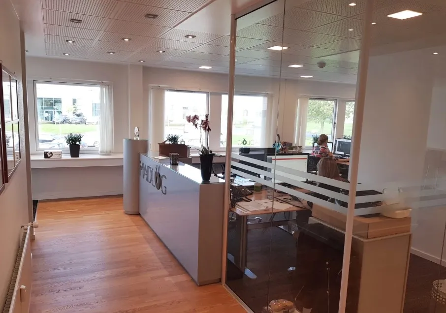 Kontorlokale med lyse gulve og store vinduer samt et moderne skrivebord med computer og blomster i en vase