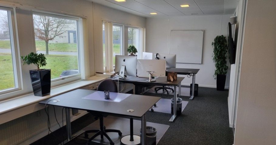 Kontor med to borde og højtalere, en whiteboard og plante samt udsigt til grønne enge og træer