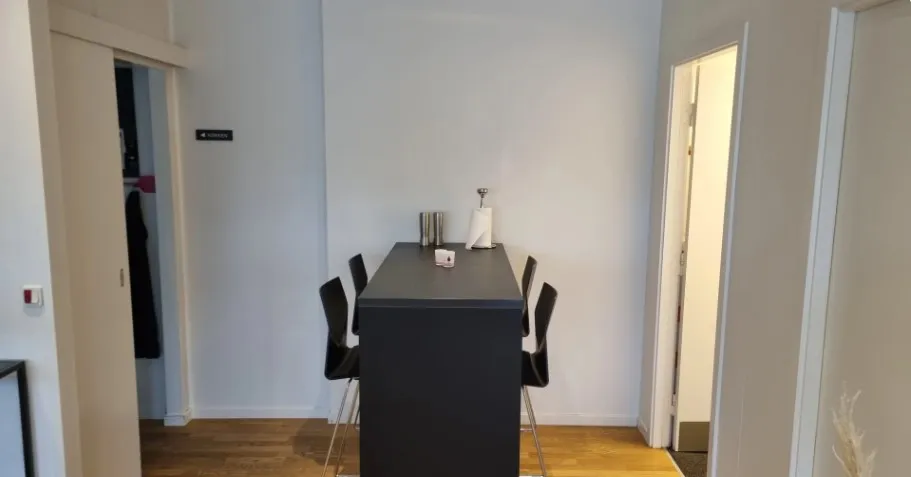 Et kontorlokale med høje hvide vægge og trægulv. Værelset har et bord med fire stole og et toilet. Der er et åbent dørkar og