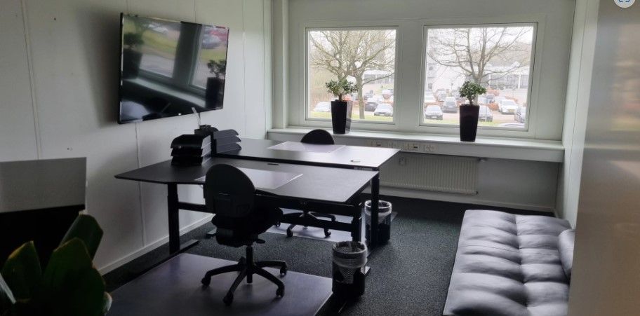 Kontorlokale med skriveborde, stole, plantepotte, tv, radiator og to vinduer med udsigt til parkeringsplads