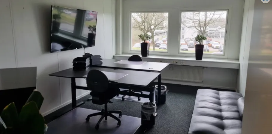 Kontorlokale med skriveborde, stole, plantepotte, tv, radiator og to vinduer med udsigt til parkeringsplads