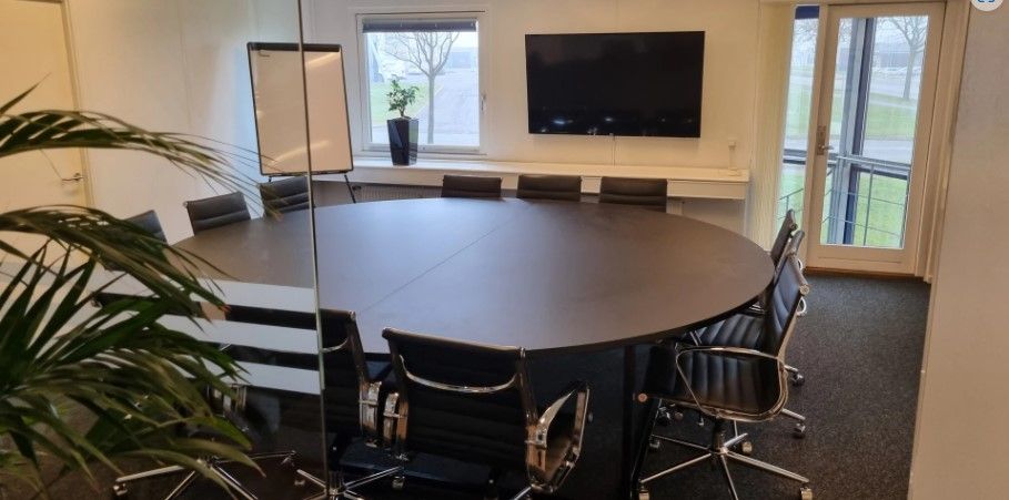 Rundt mødebord i stort kontor med moderne møbler og plante i hjørne. Tv-skærm og hvide vægge samt lyse gulv.